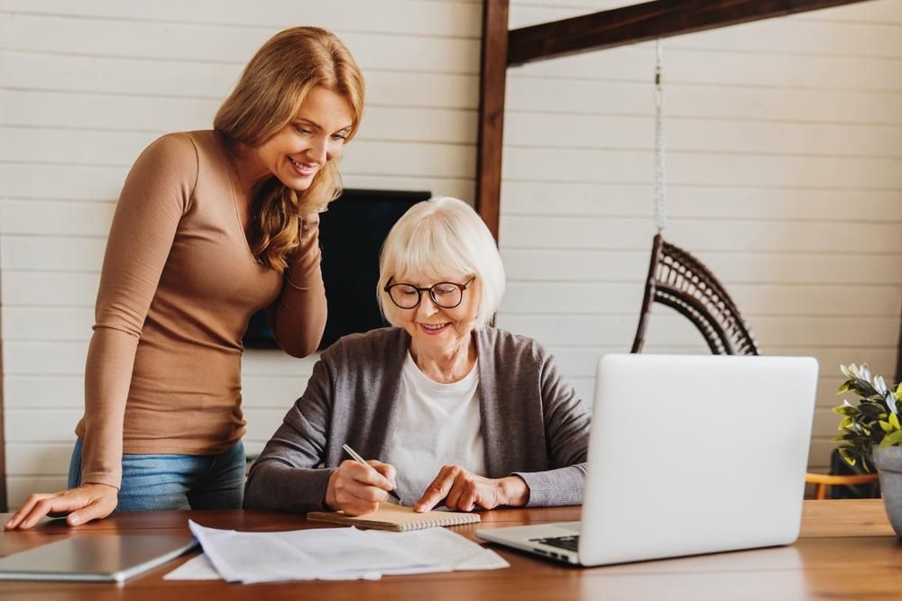Woman assisting older woman with paperwork at a table with laptop. Both smiling indoors.
