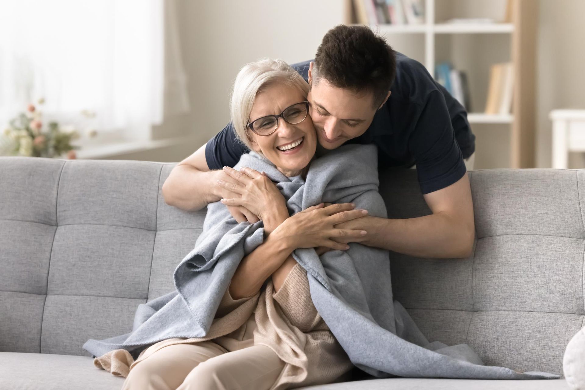 Young person hugs older person on a couch, both smiling. The older person is wrapped in a blanket.