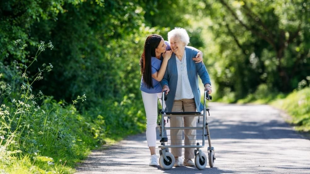 Young woman assists an elderly person using a walker on a tree-lined path. Both are smiling.