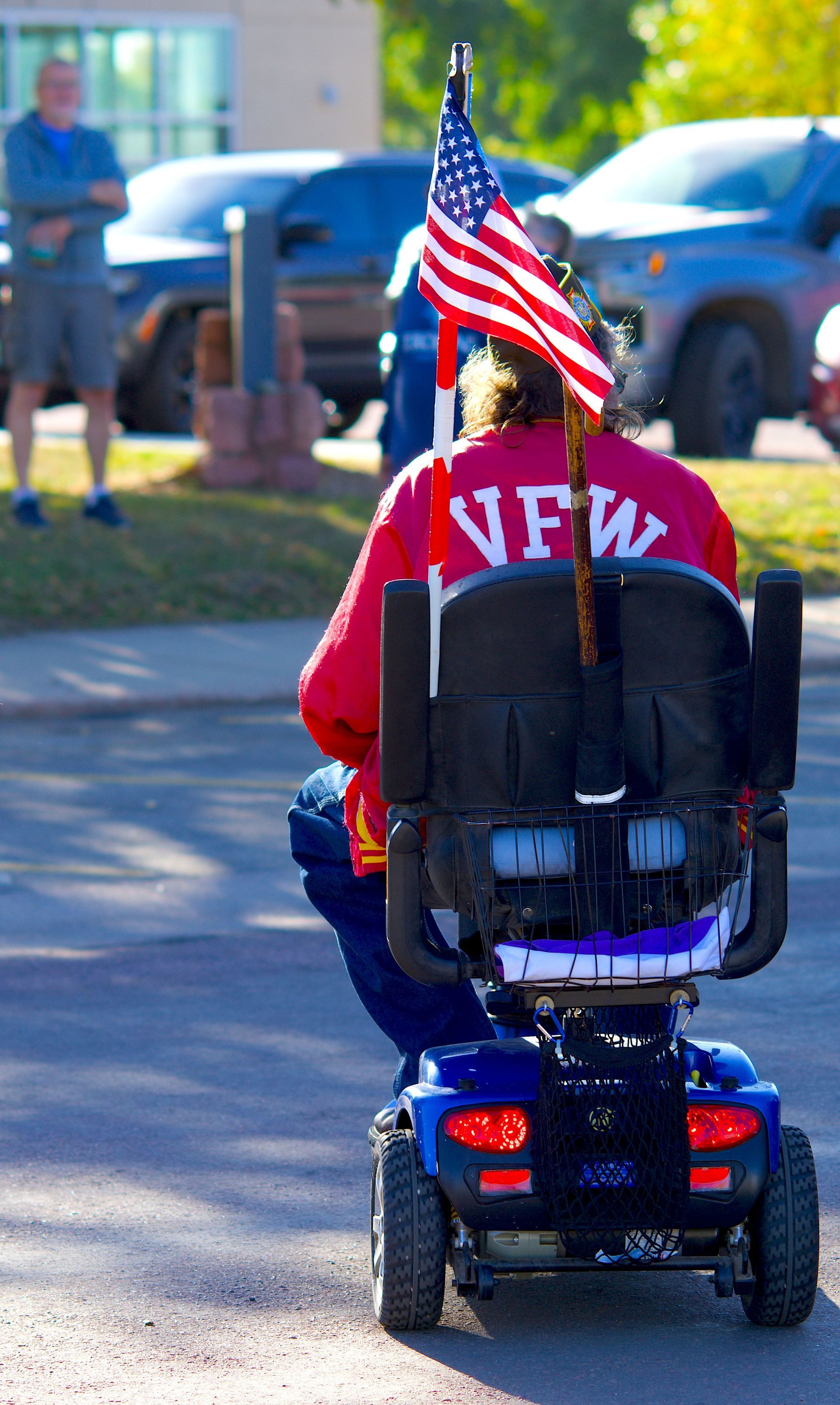 Person in a red jacket rides a mobility scooter with an American flag attached, outdoors in a parking lot.