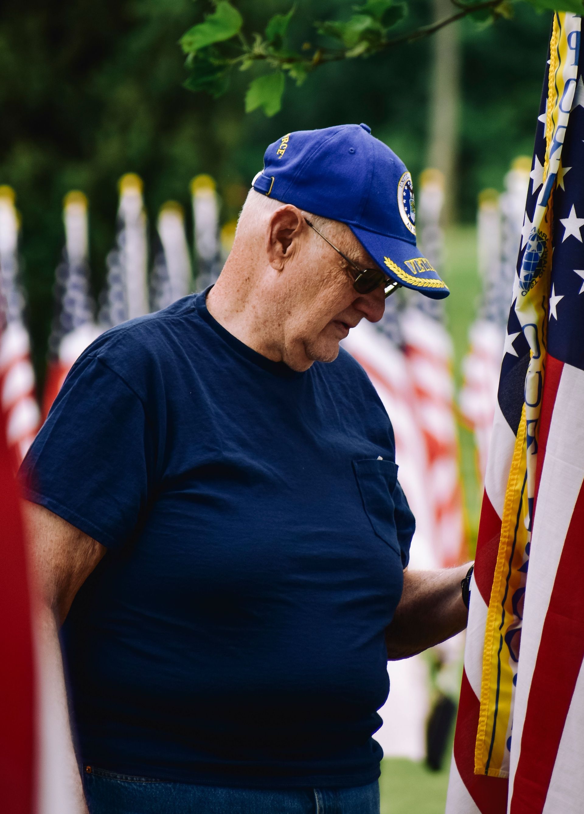 Man in blue cap and shirt standing beside a U.S. flag in a memorial setting