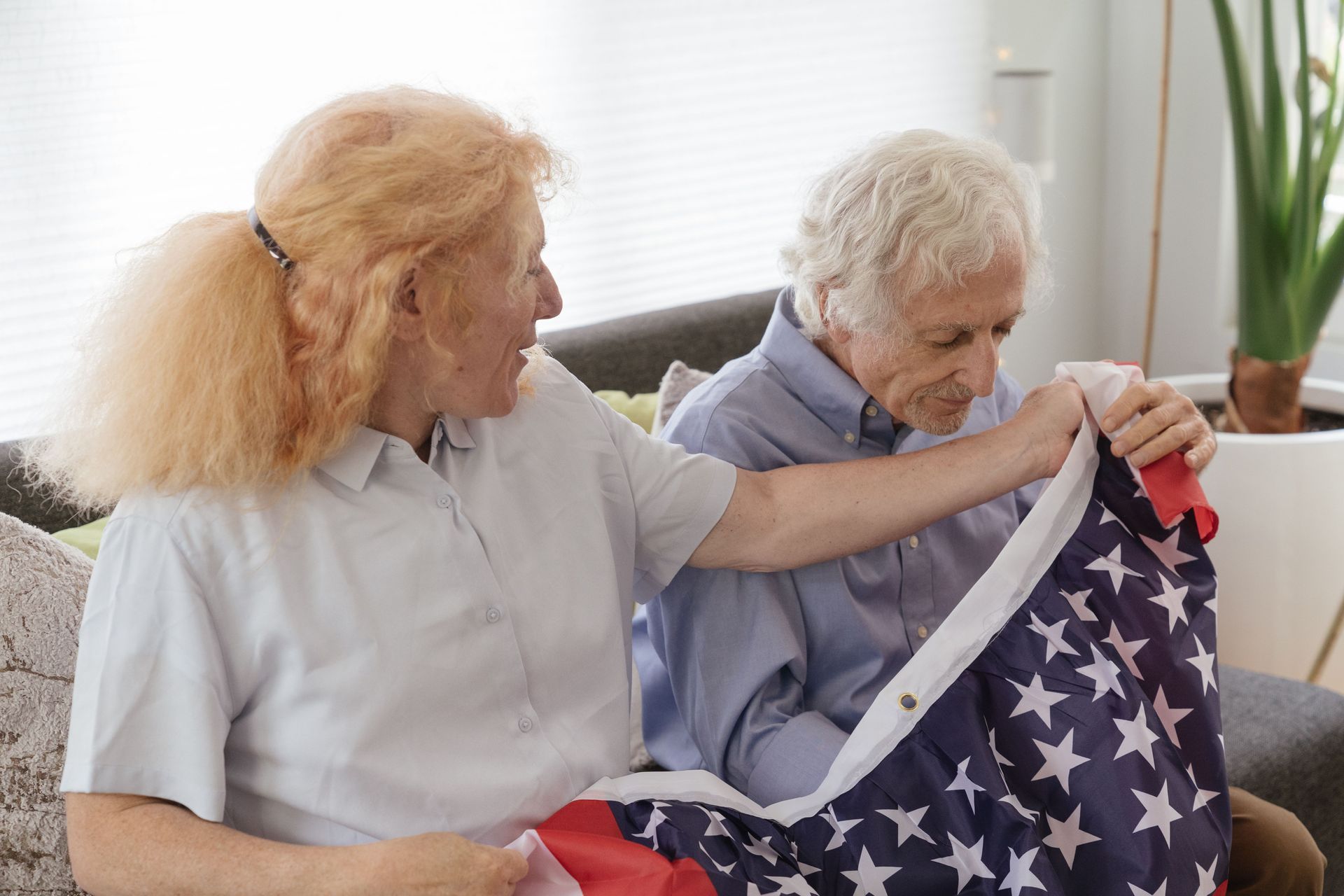 Two older people folding an American flag on a couch, with a window and plant in the background