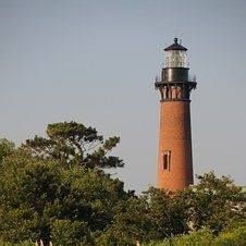Tall brick lighthouse with a black lantern above green trees under a clear sky