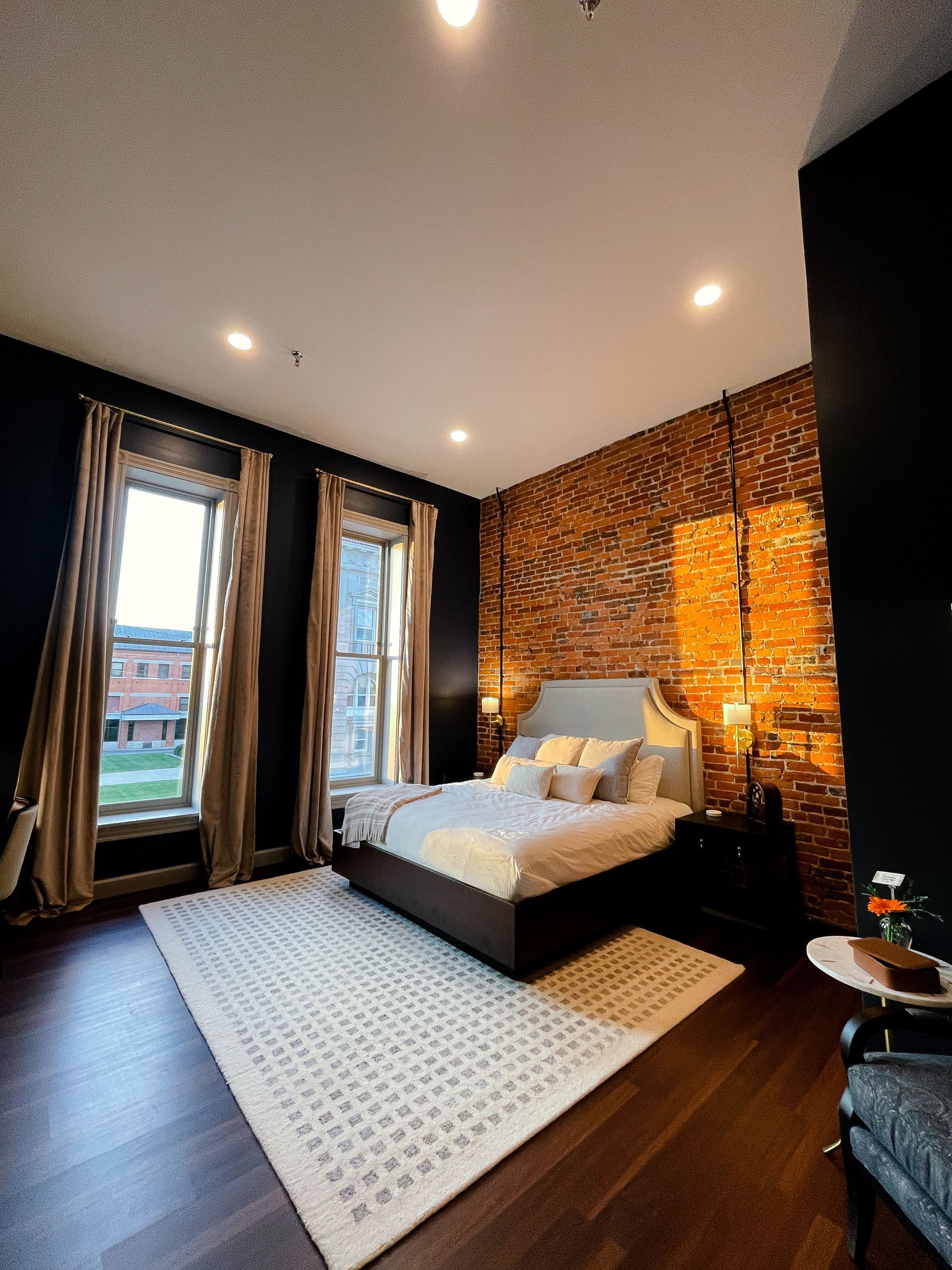 Bedroom with exposed brick wall, windows, bed, rug, and dark wood flooring.