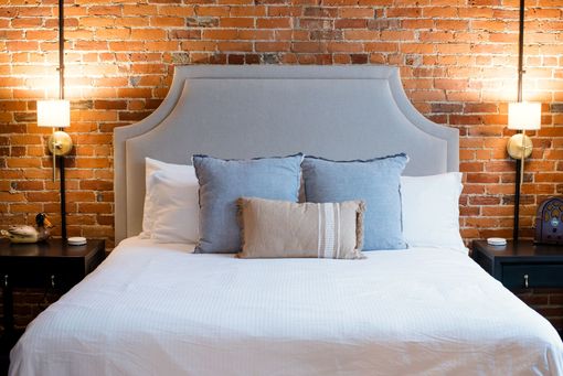 Bed with gray headboard, blue pillows, and white bedding against a brick wall; bedside lamps flank the bed.