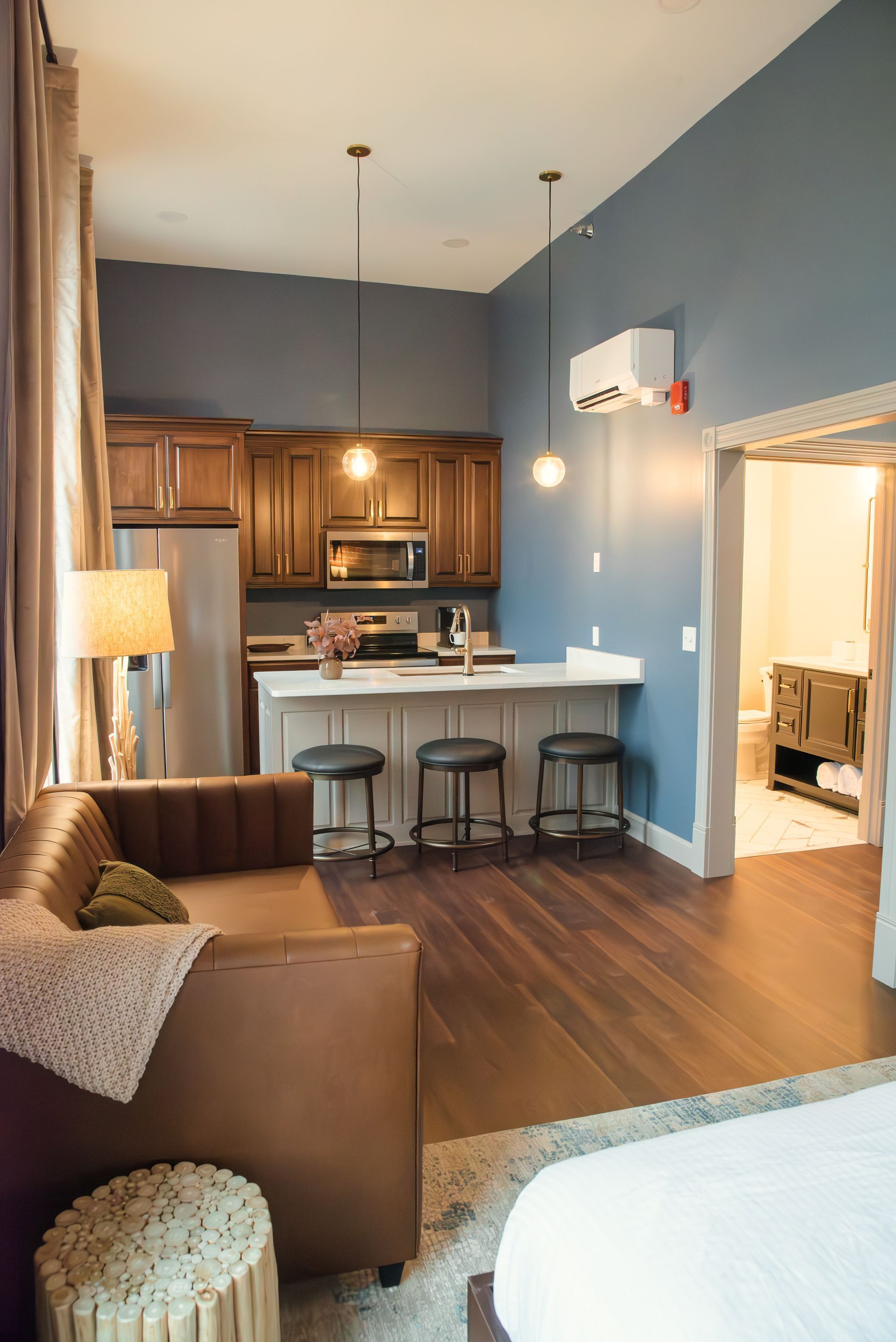 A studio apartment with a kitchen, living area, and an open doorway to a bathroom. Blue walls and wood floors.