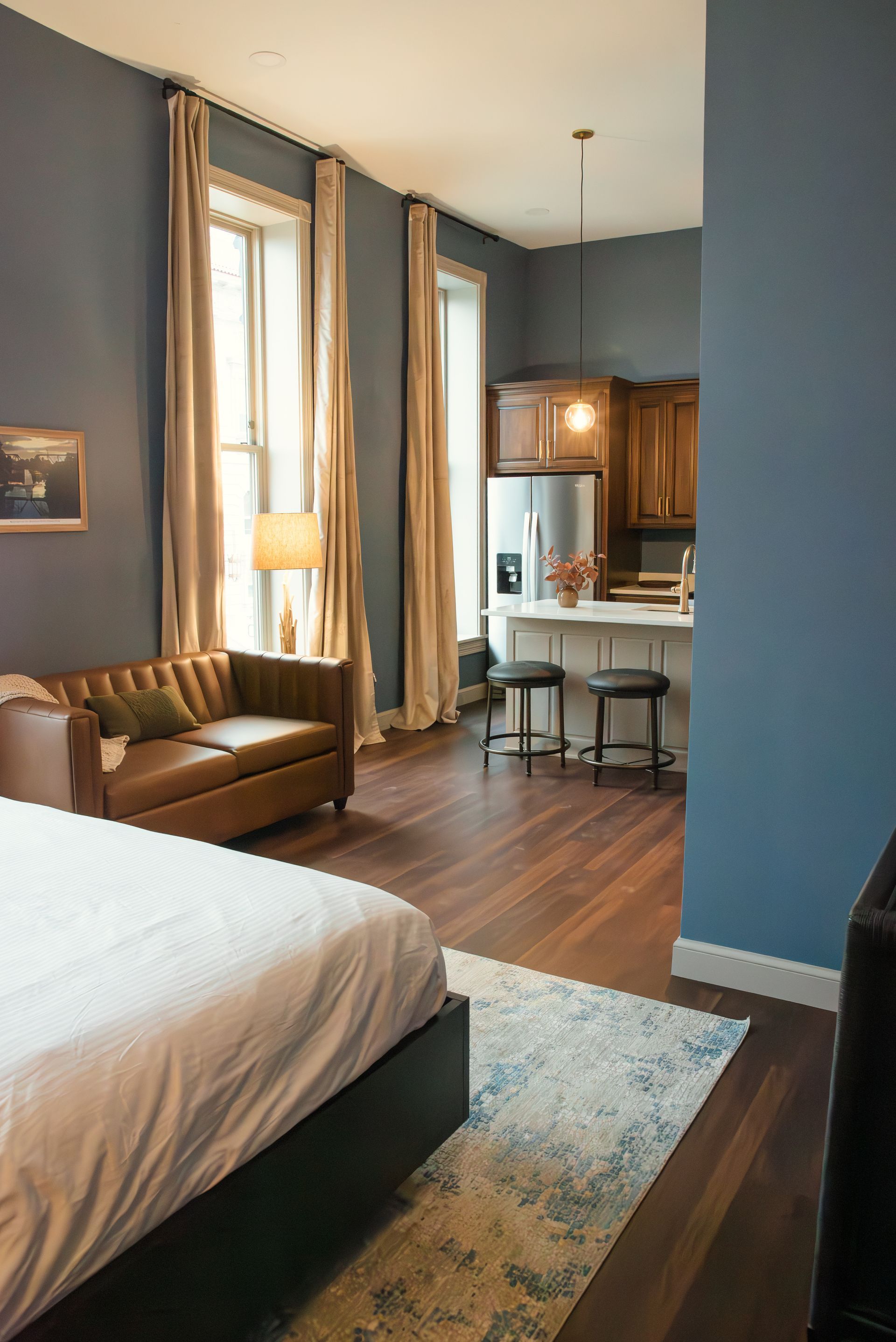 Bedroom with bed, leather sofa, kitchen. Blue walls, wood floors, beige curtains.