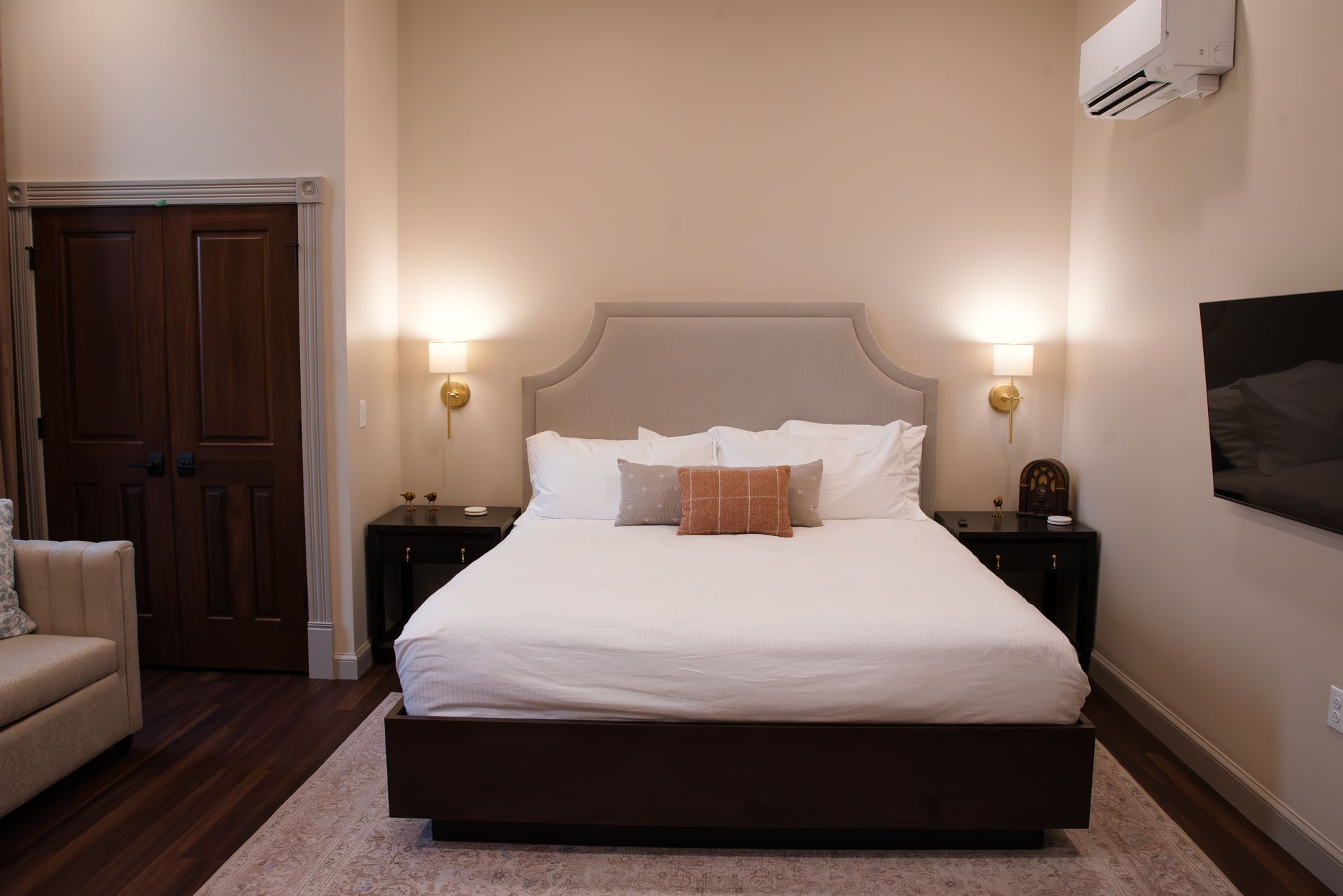 Bedroom with a queen-sized bed, nightstands, sofa, TV, and wooden door; warm lighting.
