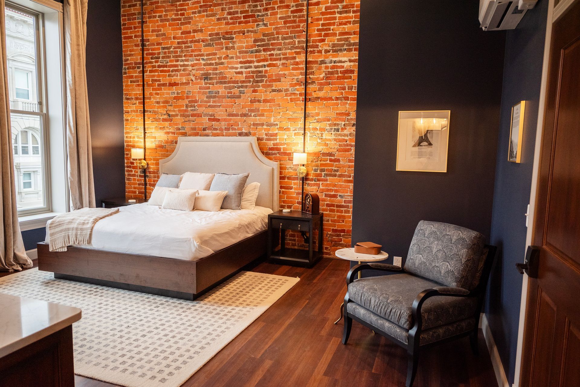 Bedroom with exposed brick wall, bed, armchair, and dark blue walls.