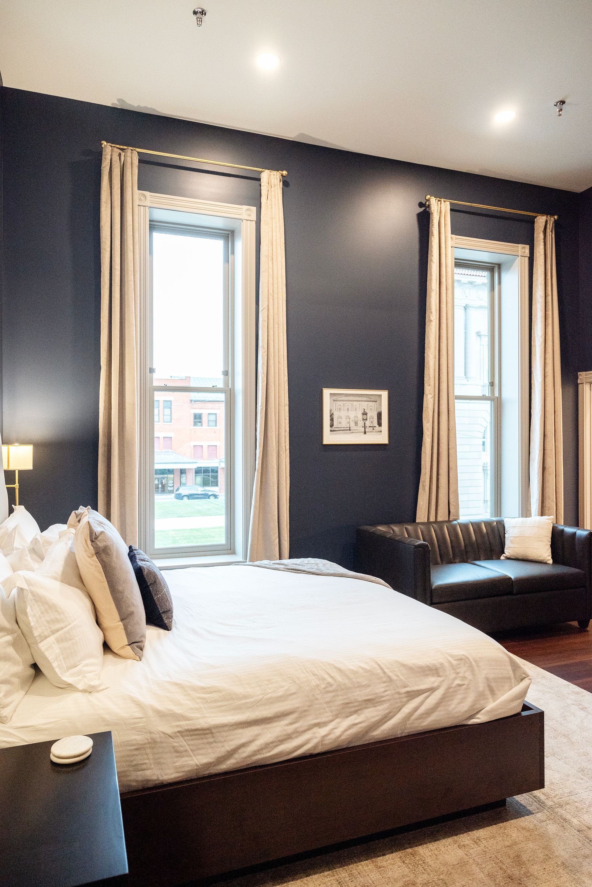 Bedroom with navy walls, large bed, sofa, and tall windows with cream curtains.