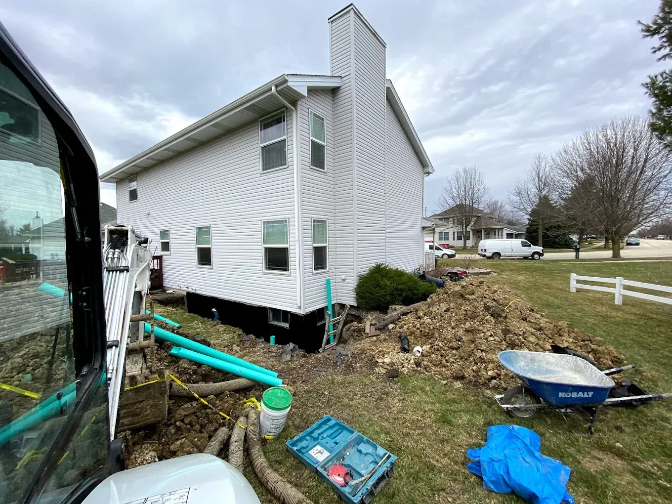 Side of a house with exposed foundation and plumbing work in progress. Dirt, tools, and a small excavator are present.