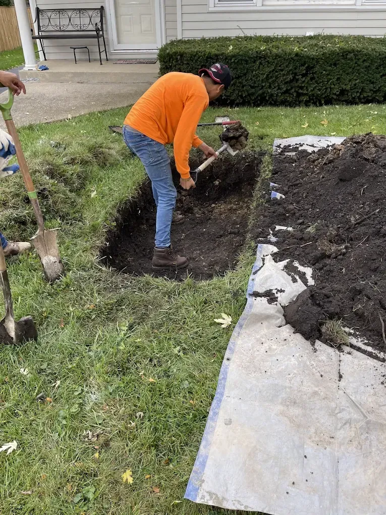 Person digging a hole in a grassy yard. Orange shirt, blue jeans, brown boots, and a black hat.