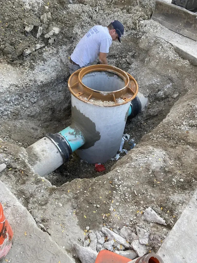 Man working on a concrete utility structure in an excavated area, with piping.