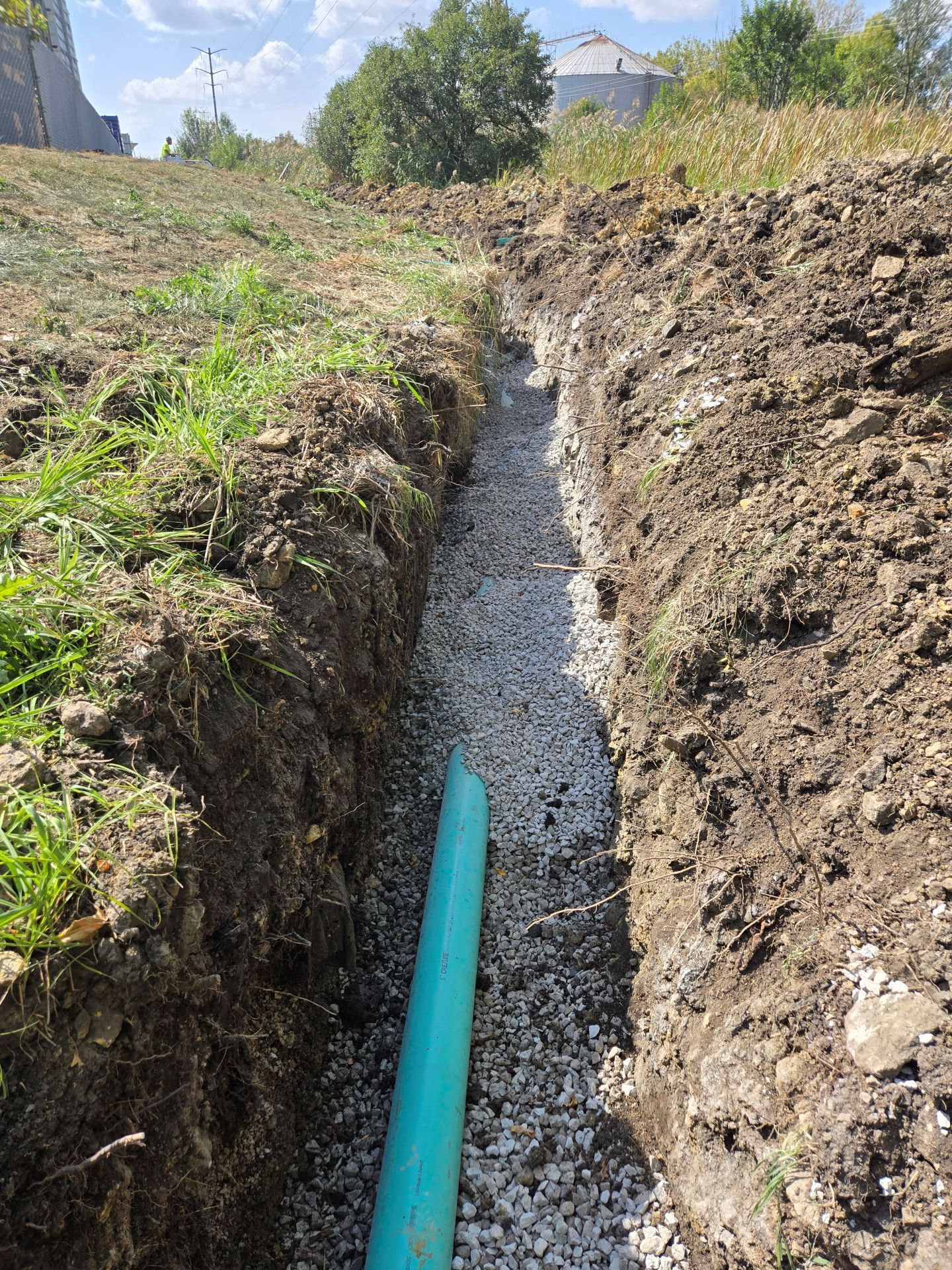 Trench filled with gravel and a blue pipe, dug into a grassy hill.