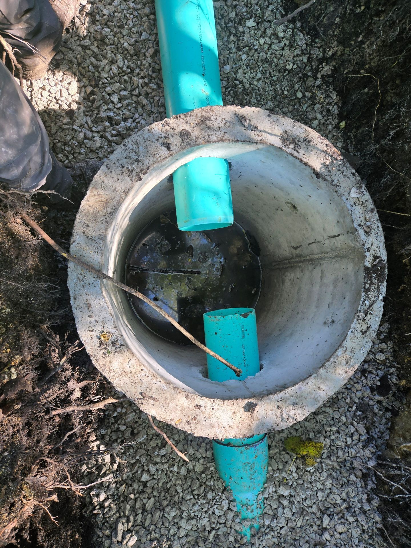 Concrete cylindrical structure with blue pipes, set in gravel ground.