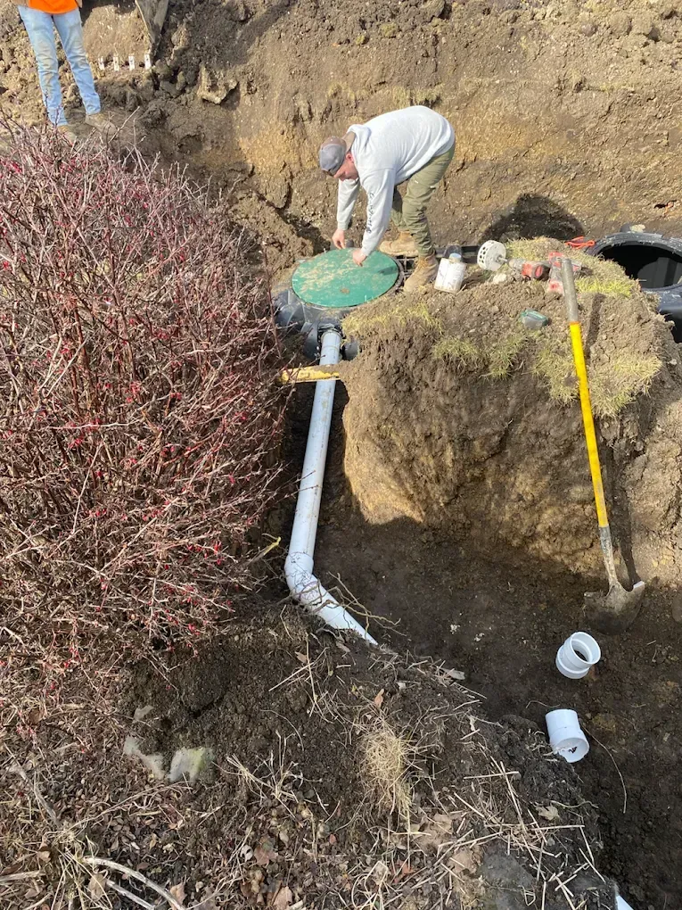 Workers installing drainage system in a ditch; white pipe, dirt, and shovel present.