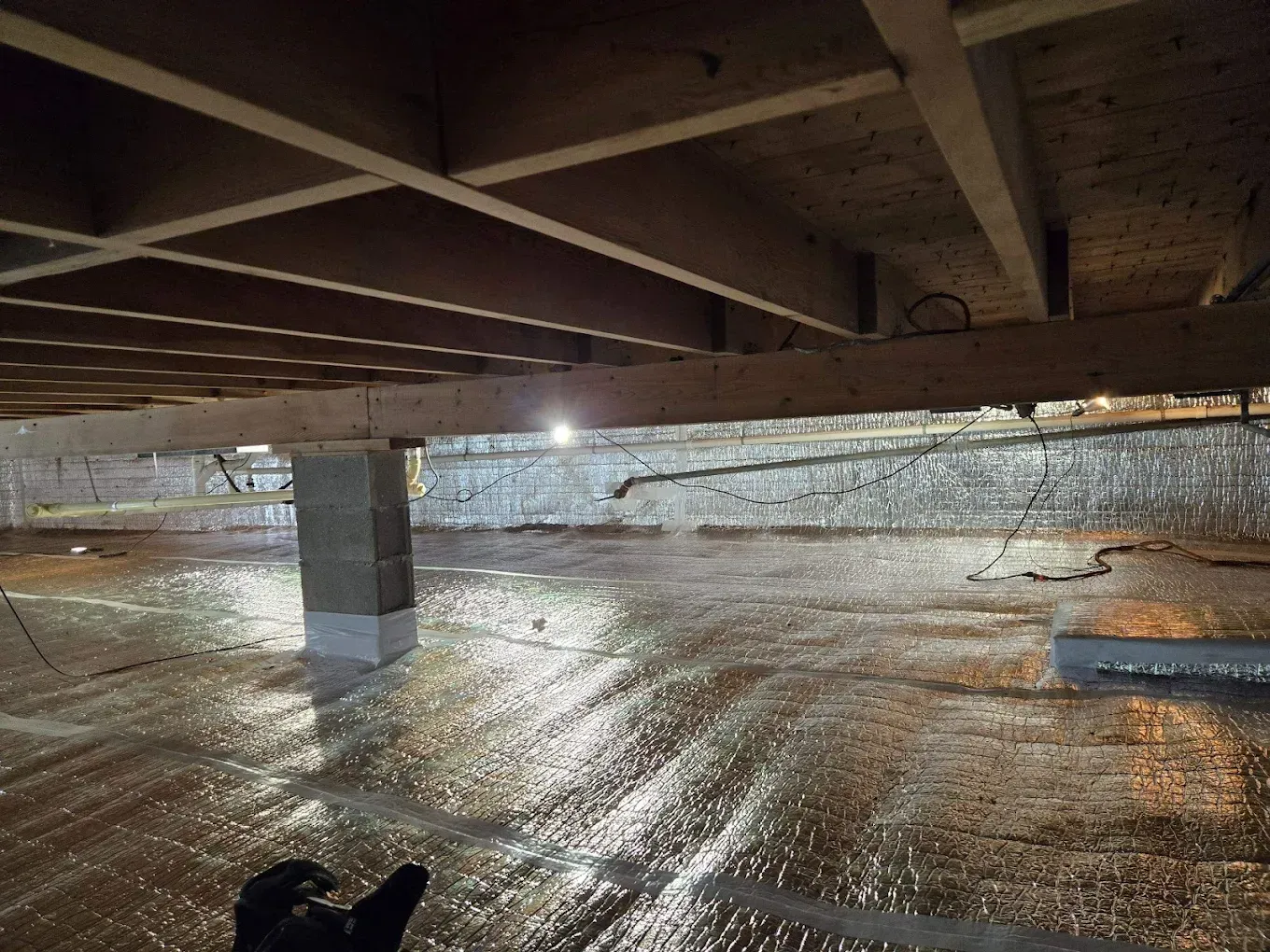 Crawlspace with insulation and support beams. Reflective material covers the ground. Wood joists above.