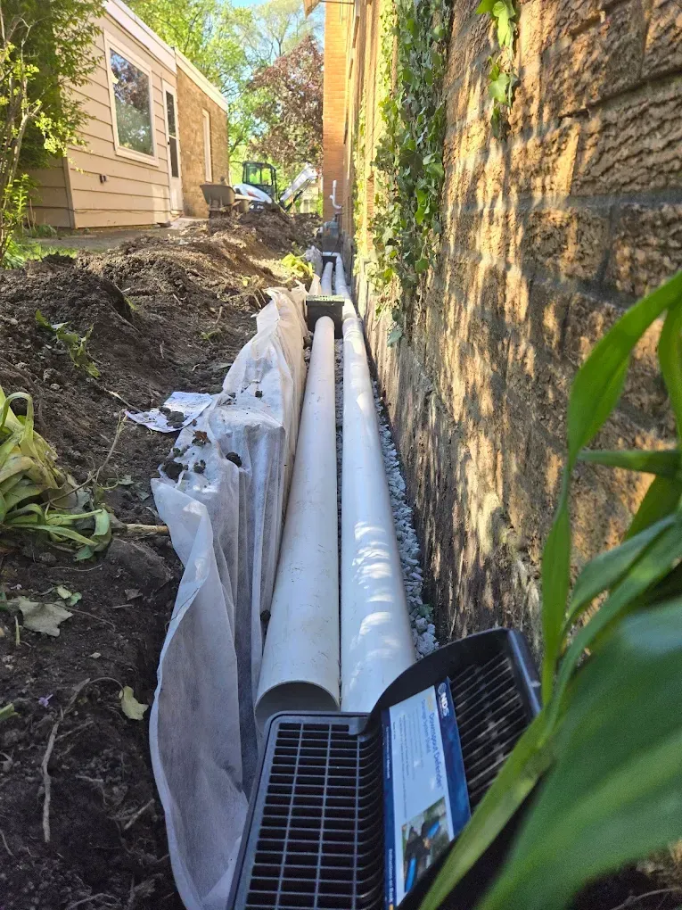 A trench with drain pipes next to a stone wall. White landscape fabric and a grate are visible.