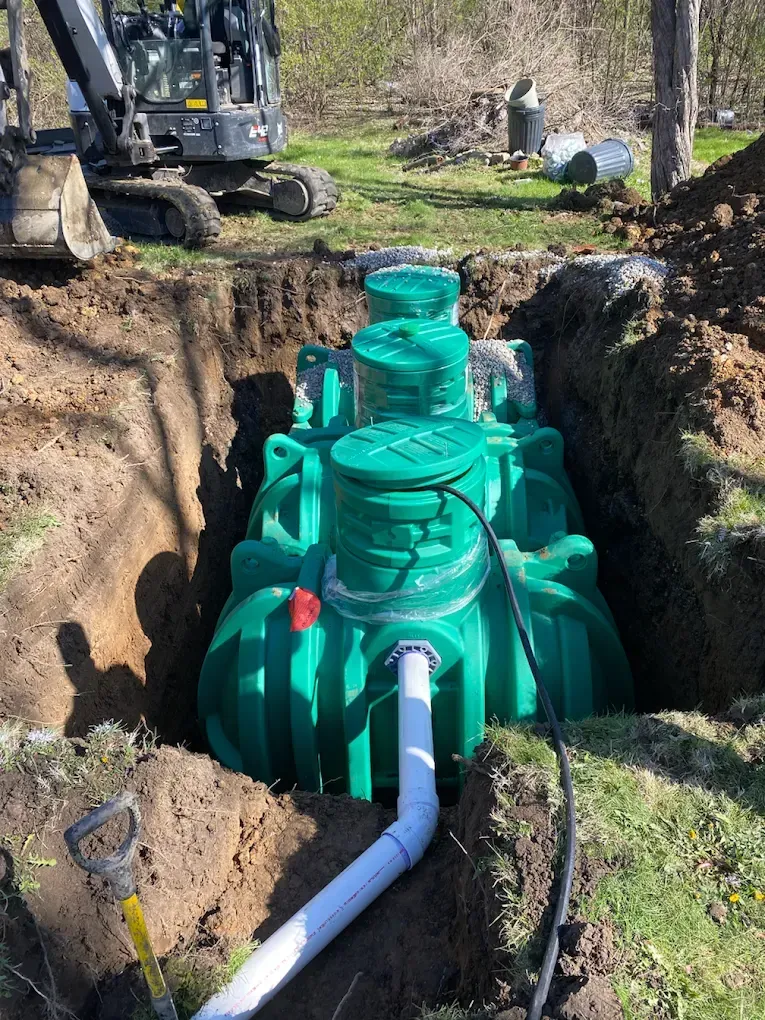 Green septic tank installed in a dug pit, with a white pipe and an excavator nearby.