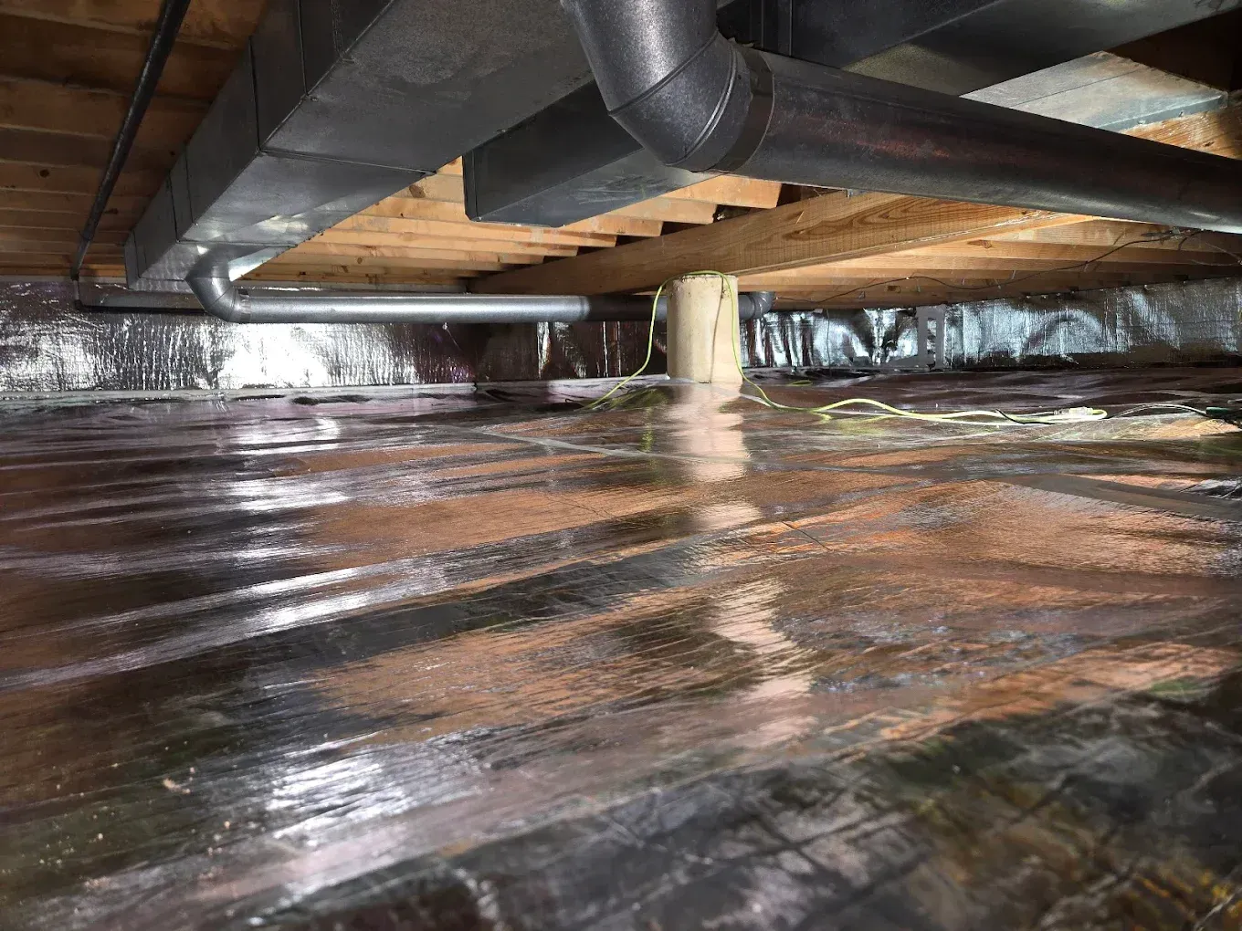 View of a crawl space with pipes, wooden beams, insulation, and a reflective barrier.