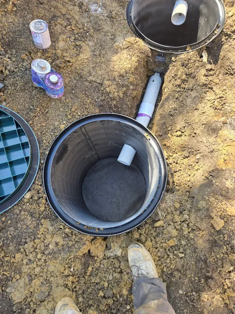 Open septic tank with pipe connections, a ground-level view. Earth and construction materials surround.