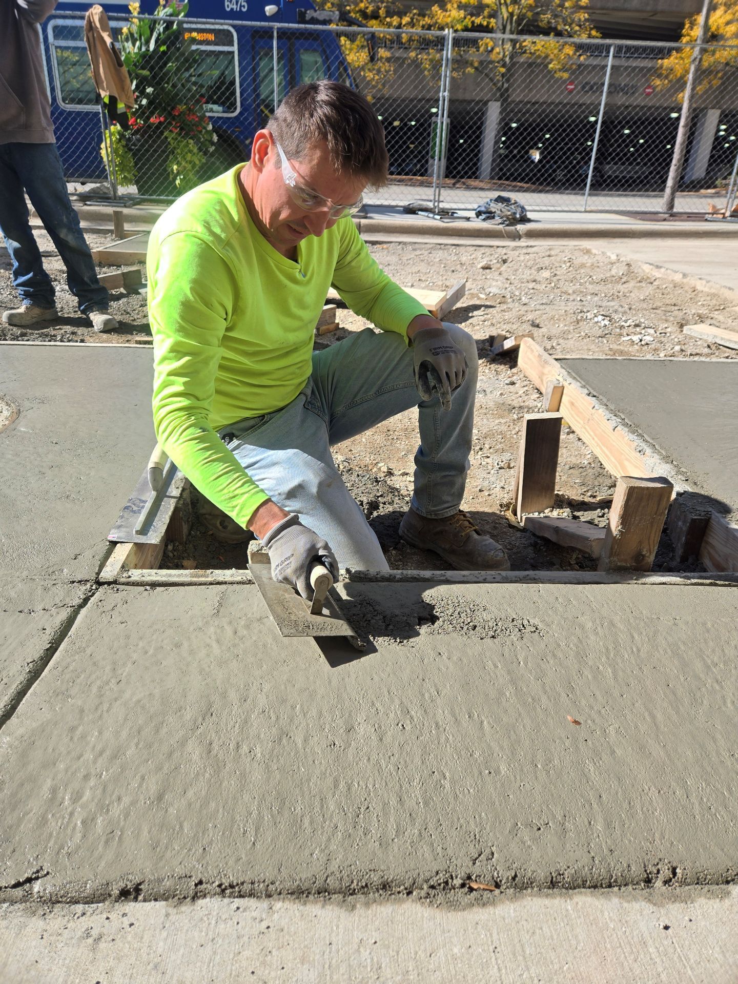 Construction worker smoothing concrete with a trowel outdoors. He wears a yellow shirt, jeans, and gloves.