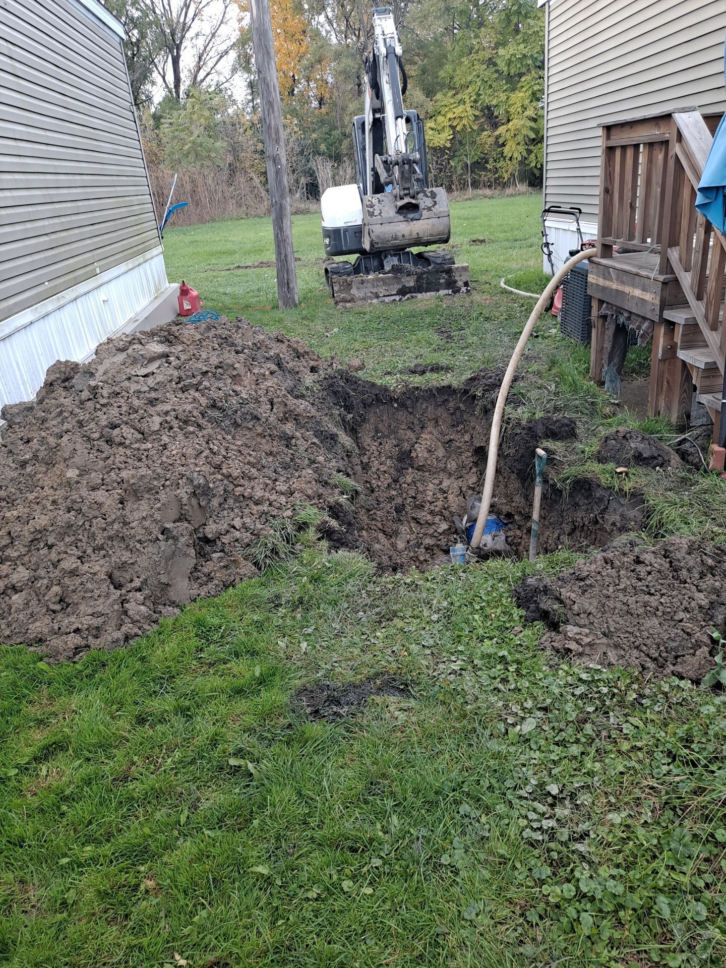 A backhoe digs a large hole in a grassy yard, near a house and a pile of dirt.