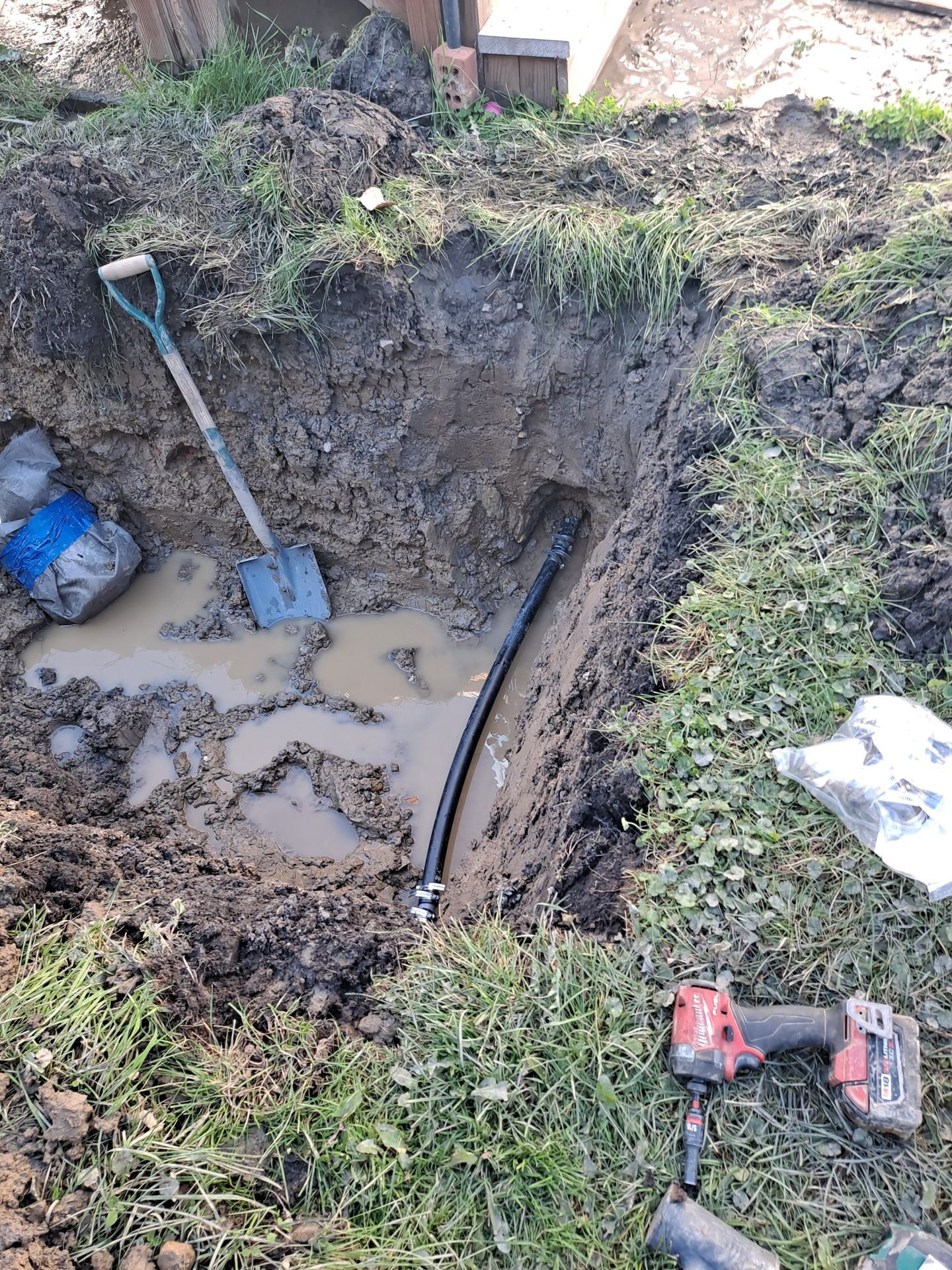 Dug out trench with black pipe in wet soil, shovel, and power drill on grass.