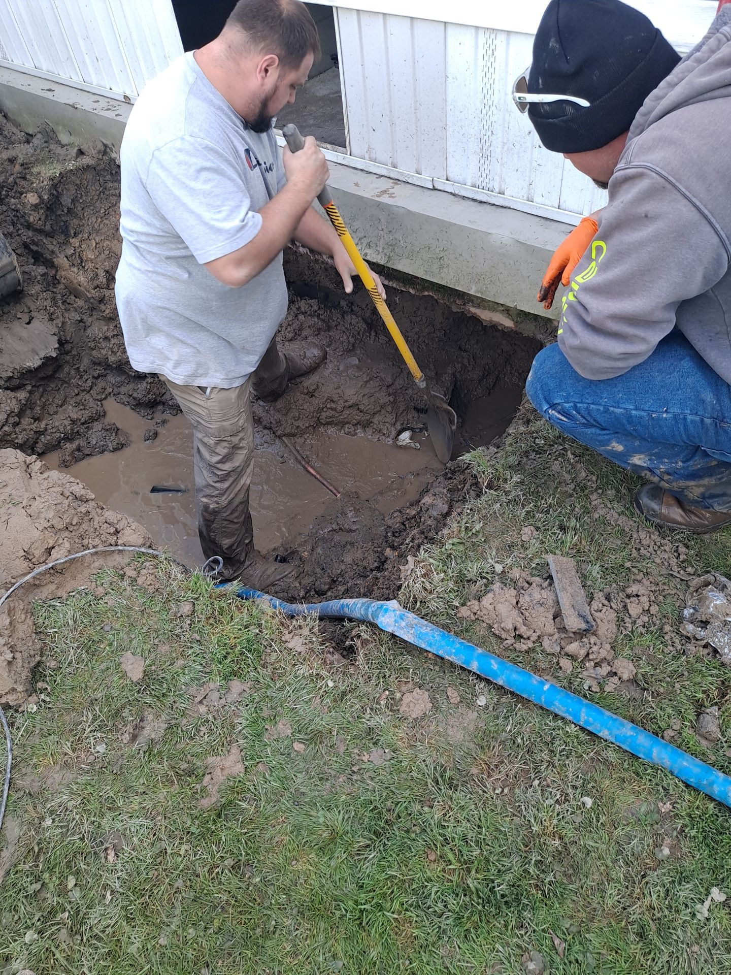 Two men dig in muddy earth near a building. One uses a shovel, the other kneels. A blue hose lies on grass.