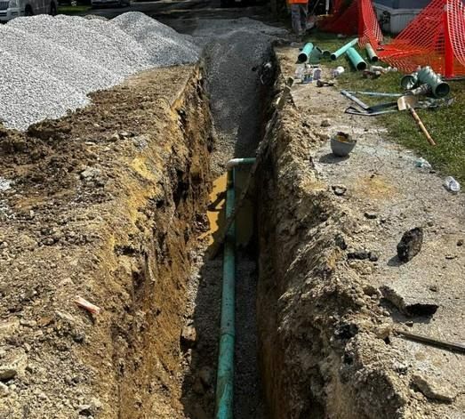 Trench dug in soil containing a green pipe, with gravel, pipes, and safety cones nearby.