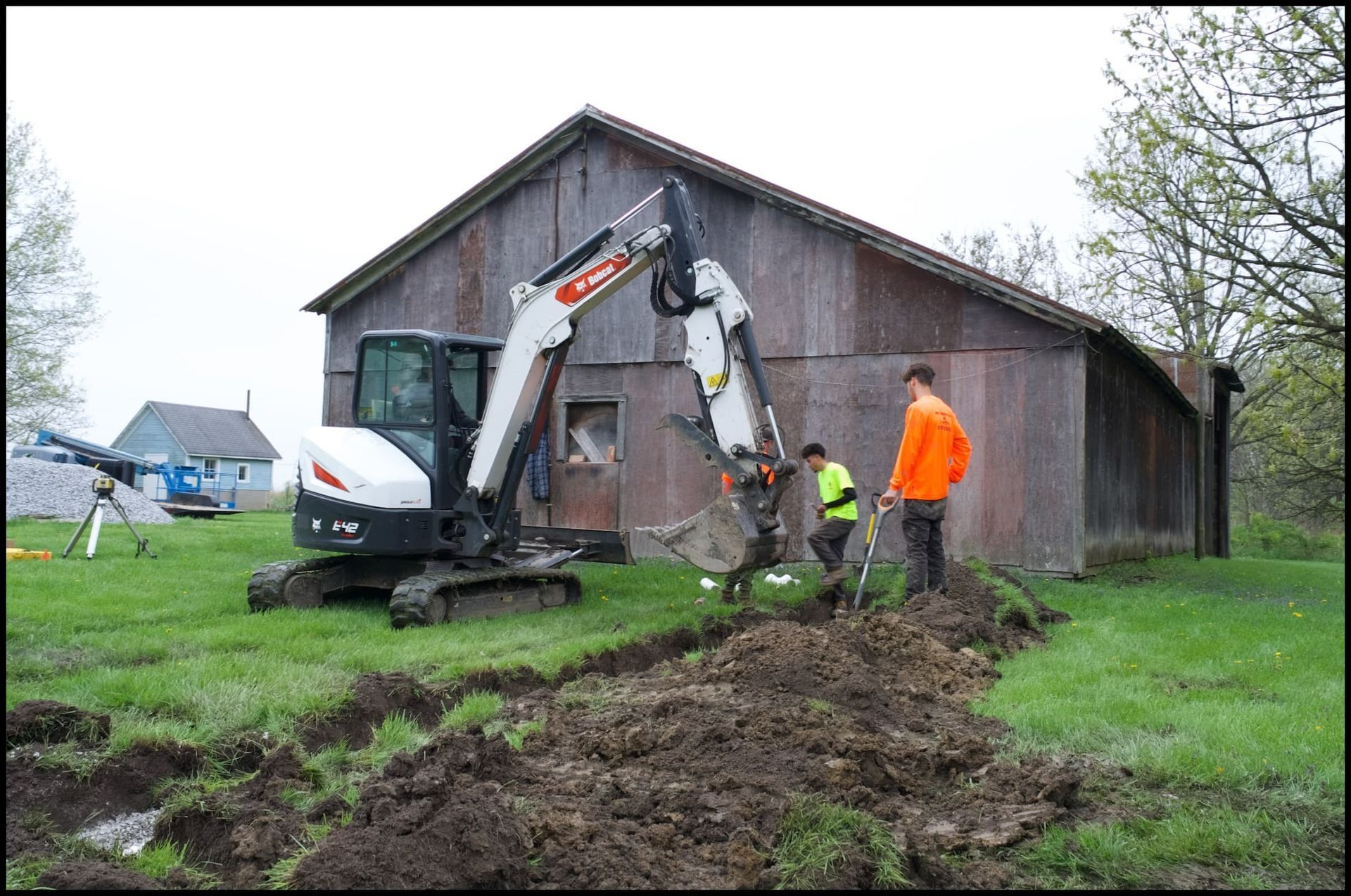 Bobcat excavator digging trench near weathered barn; two workers in orange vests.