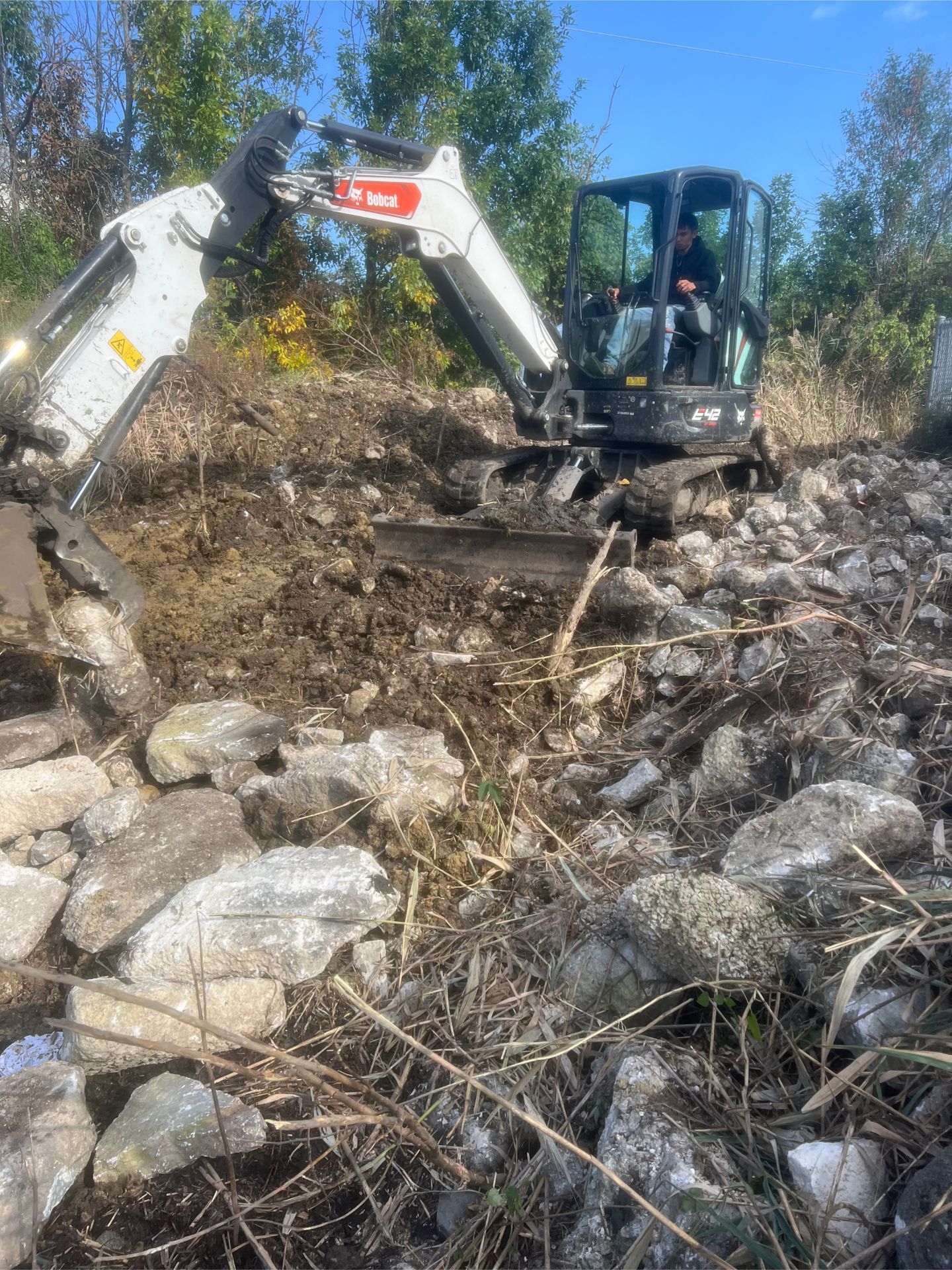 A small Bobcat excavator digs into a rocky hillside. A person sits in the cab operating the machine.