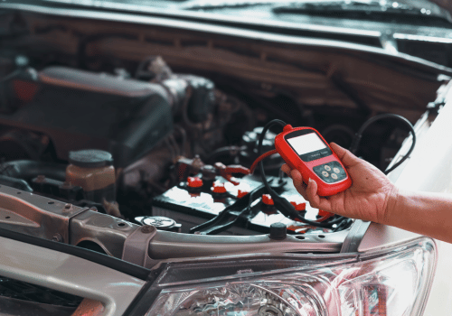 Hand holding automotive diagnostic scanner while testing vehicle battery and electrical system under