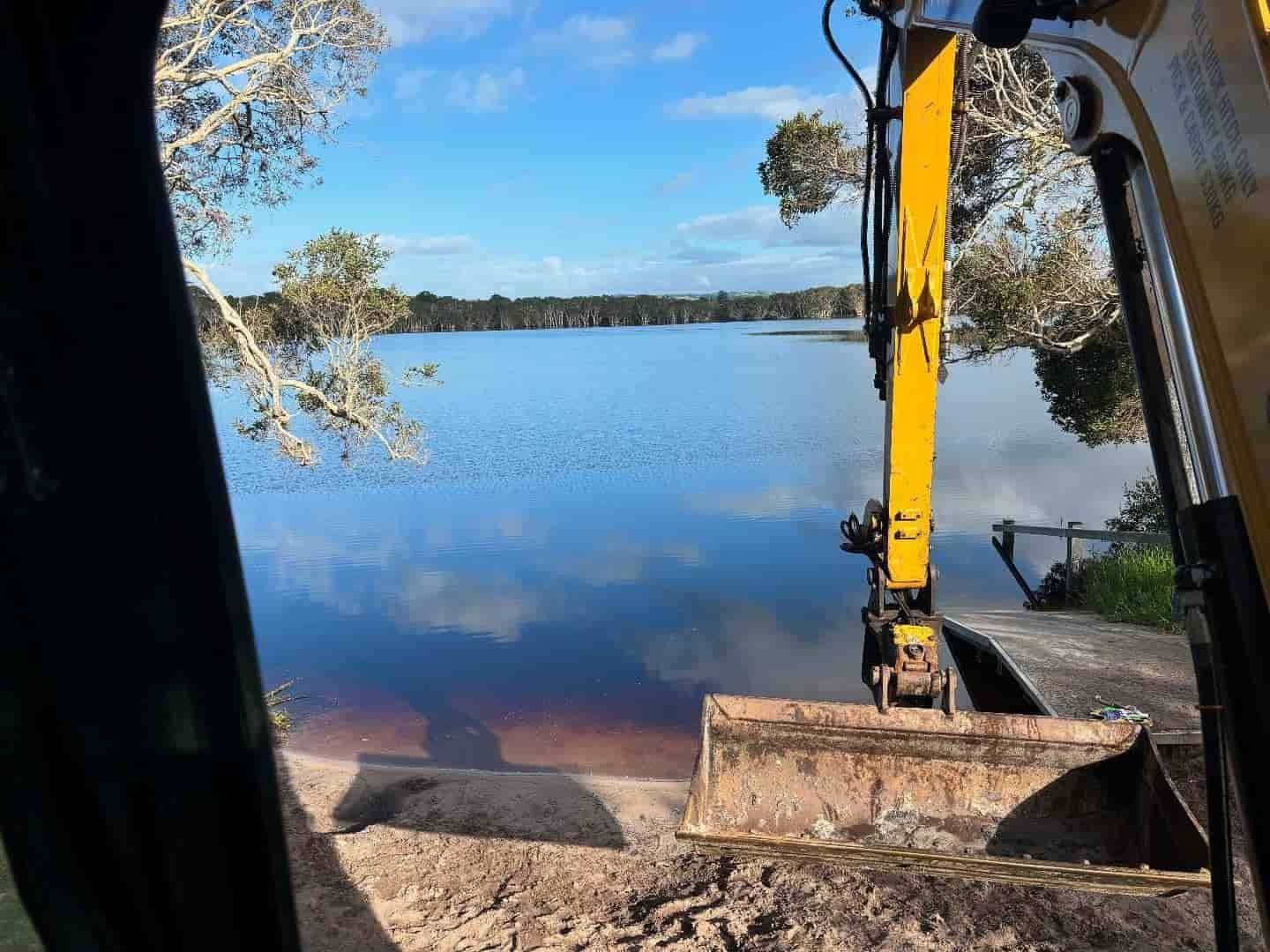 A yellow excavator scoops earth from a bank into a still, blue lake reflecting the sky.