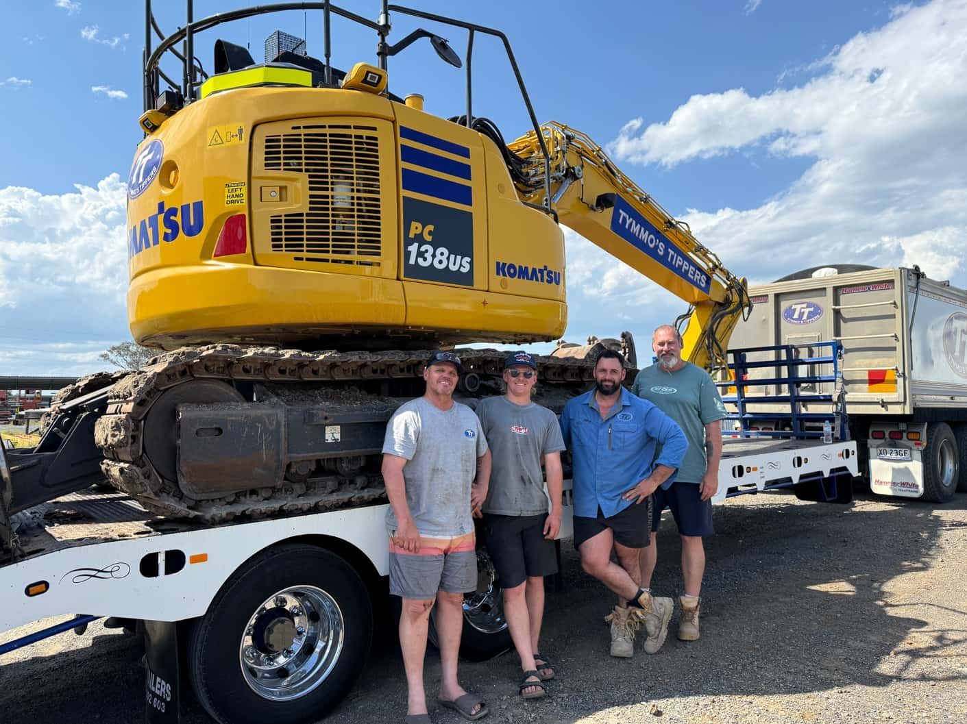 Four people stand in front of a yellow Komatsu excavator on a trailer, under a blue sky.