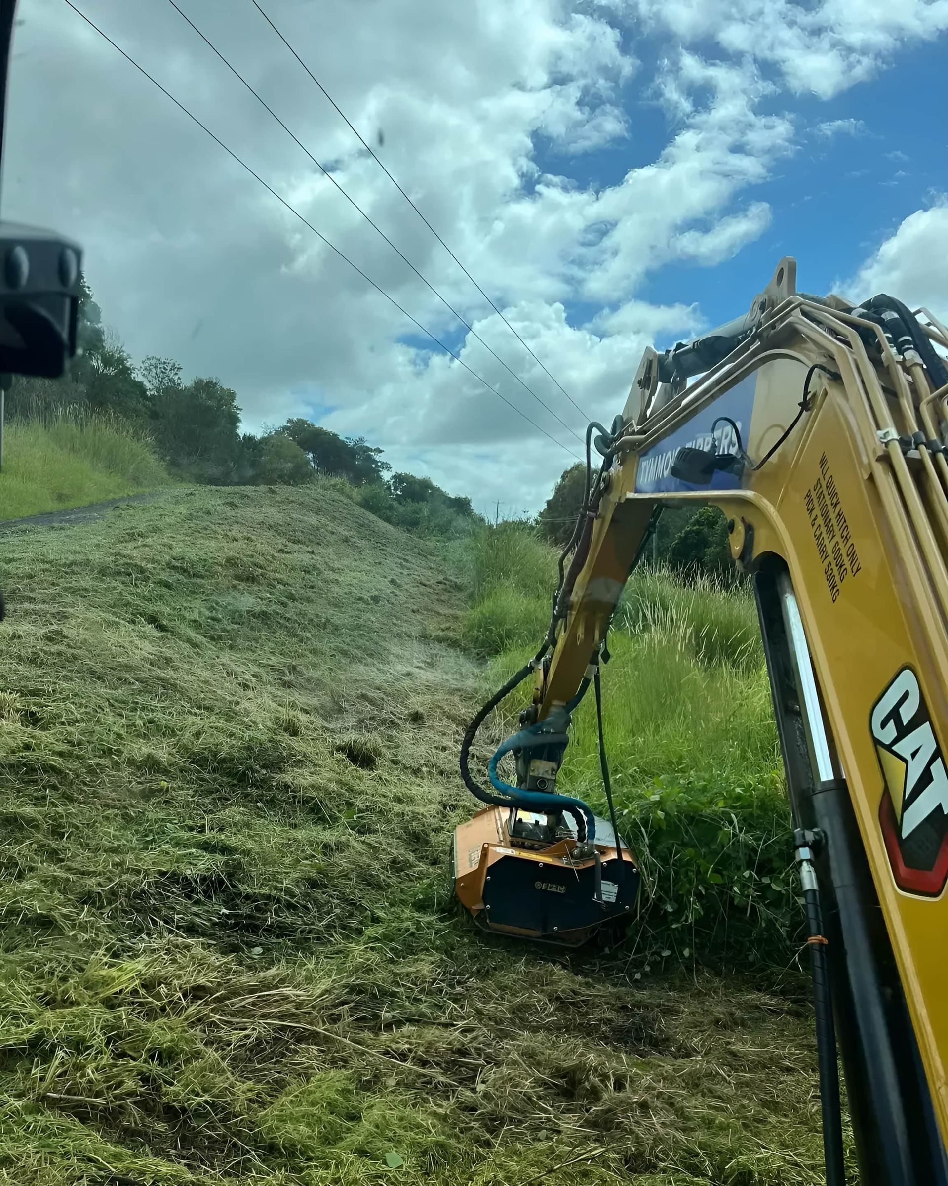 A yellow CAT excavator with a brush cutter clearing vegetation on a hillside under a cloudy sky.