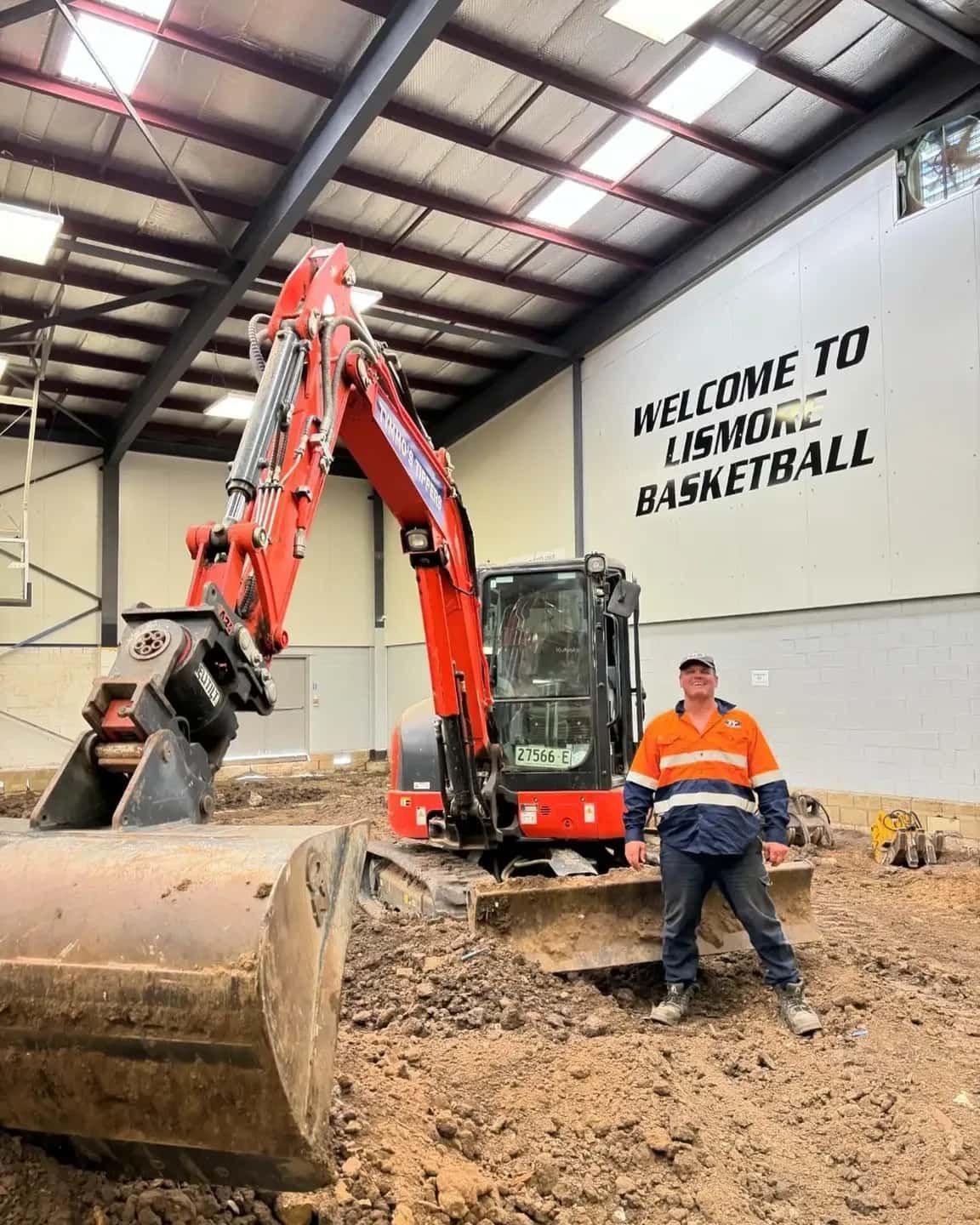 A Man Is Standing In Front Of An Excavator In A Warehouse — Tymmo's Tippers In Lennox Head, NSW