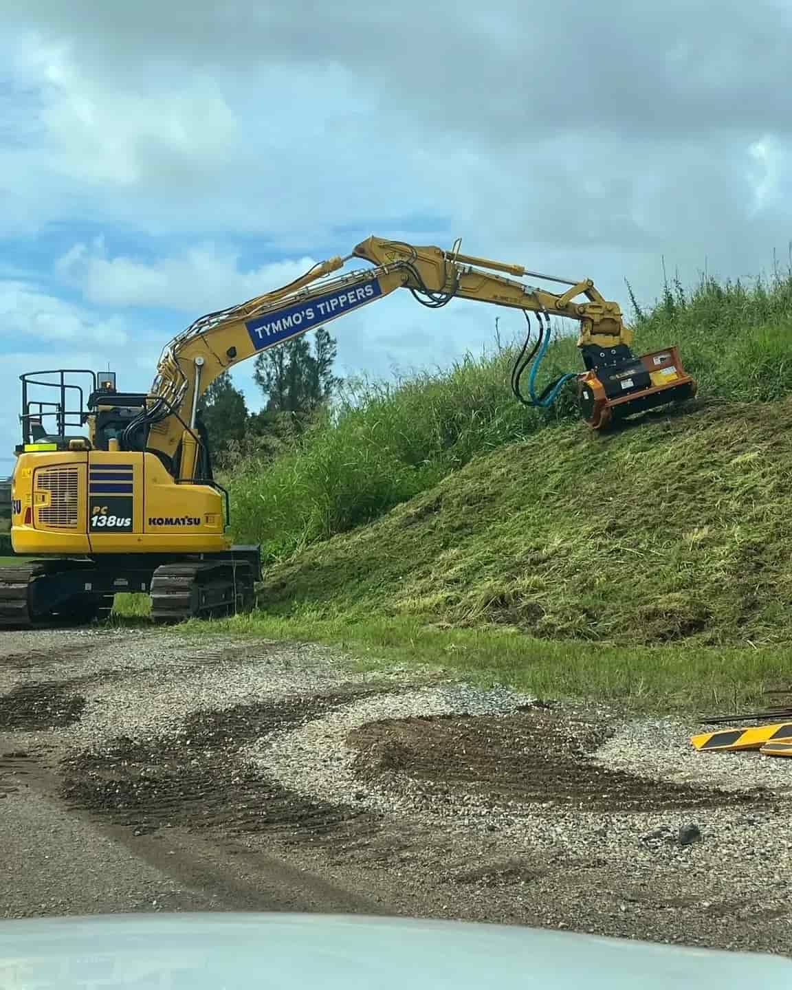 A Yellow Excavator Is Cutting Grass On A Hill — Tymmo's Tippers In West Ballina, NSW