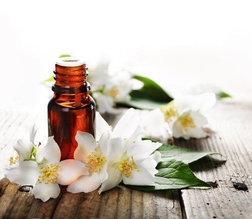 Flacone marrone di olio essenziale circondato da fiori di gelsomino bianchi su una superficie di legno.