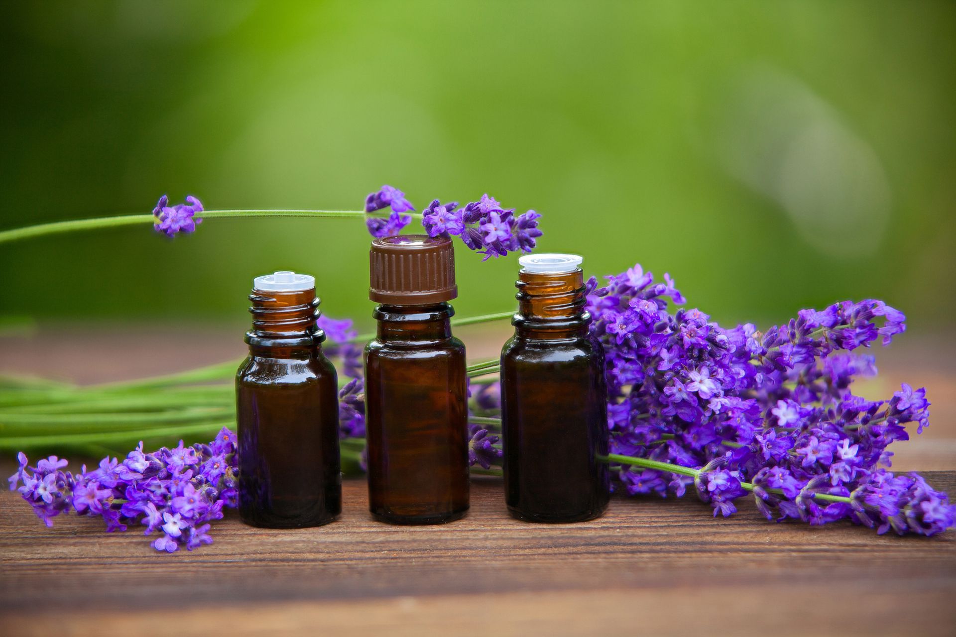 Tre bottiglie di olio essenziale marrone con fiori di lavanda su una superficie di legno.