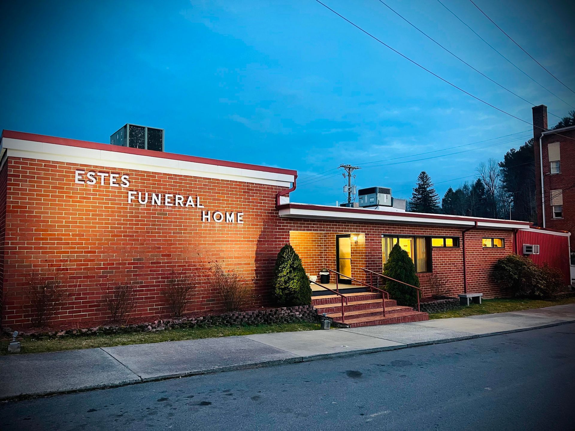 Estes Funeral Home: A one-story red brick building with lit entrance and windows under a twilight blue sky.