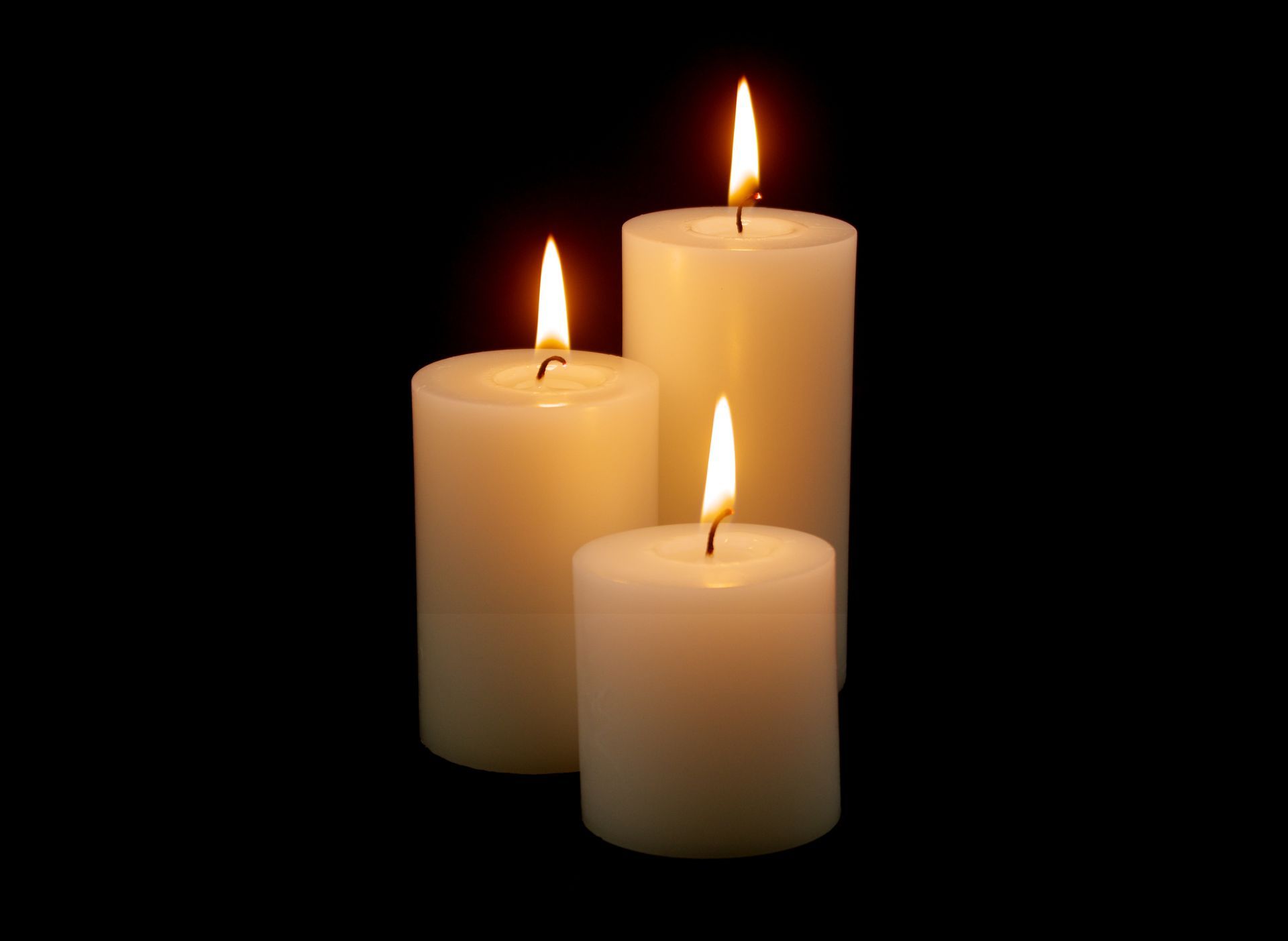 Three off-white pillar candles of varying heights, each with a flickering flame, set against a solid black background.