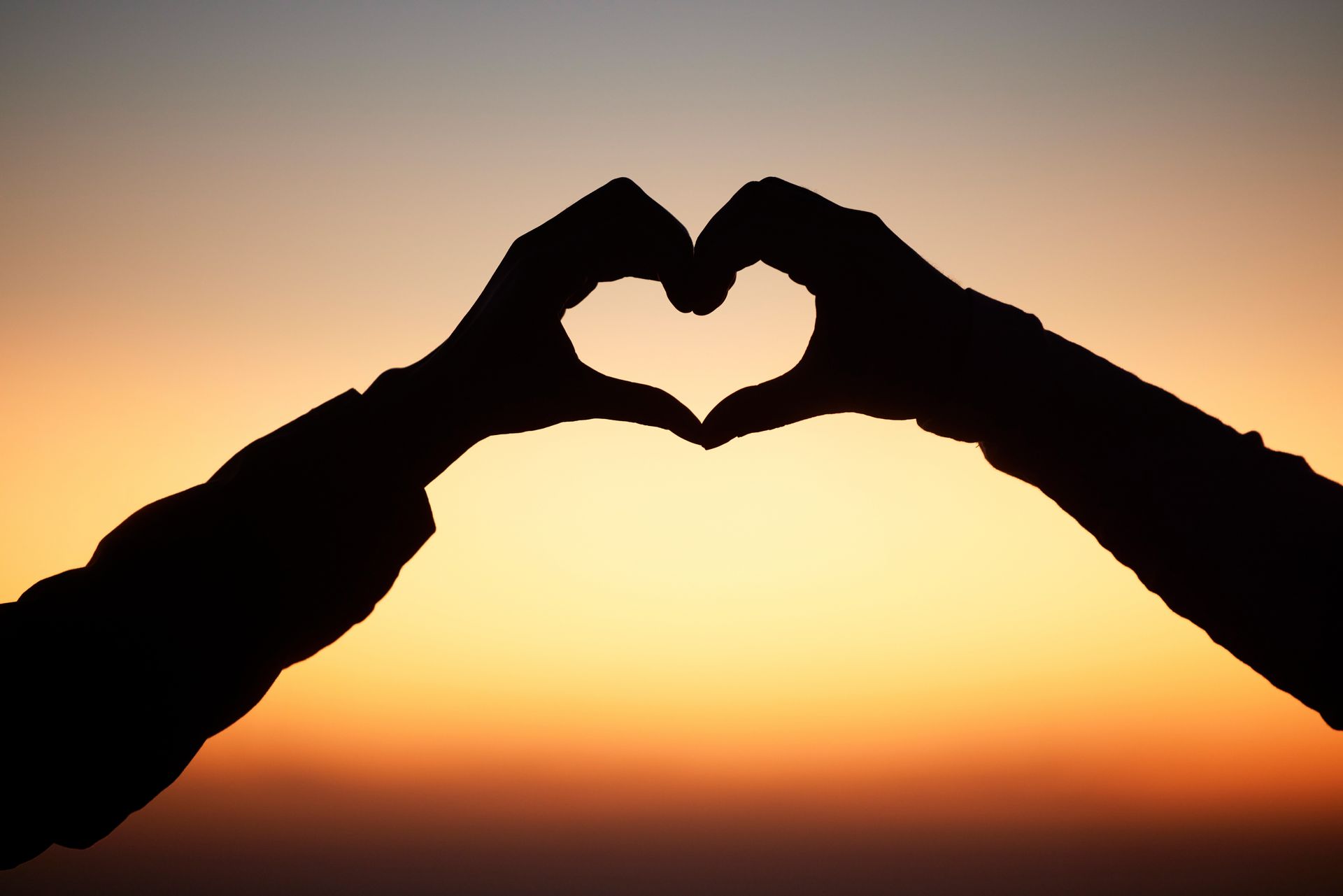 Silhouetted hands form a heart shape against a warm, orange-hued sunset sky.