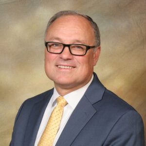 Professional headshot of a man in glasses wearing a navy suit and yellow tie against a neutral studio backdrop