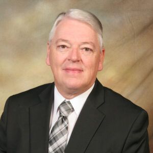 Formal studio portrait of a man in a dark suit and plaid tie against a beige background