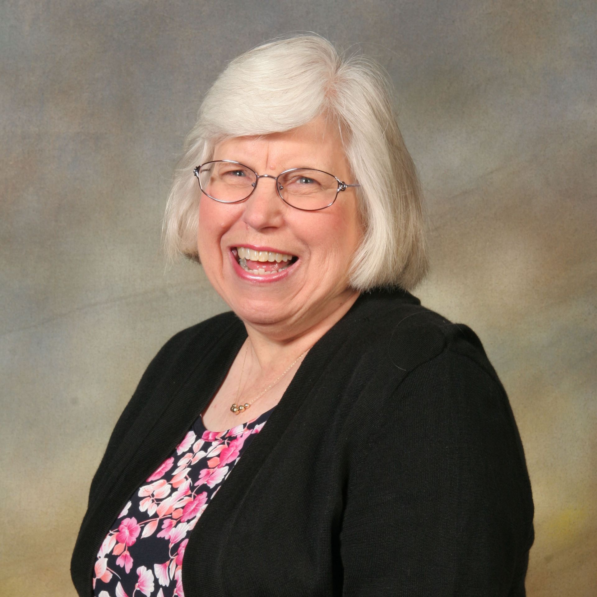 Smiling woman with white hair and glasses in a black cardigan over a pink floral top, against a studio backdrop