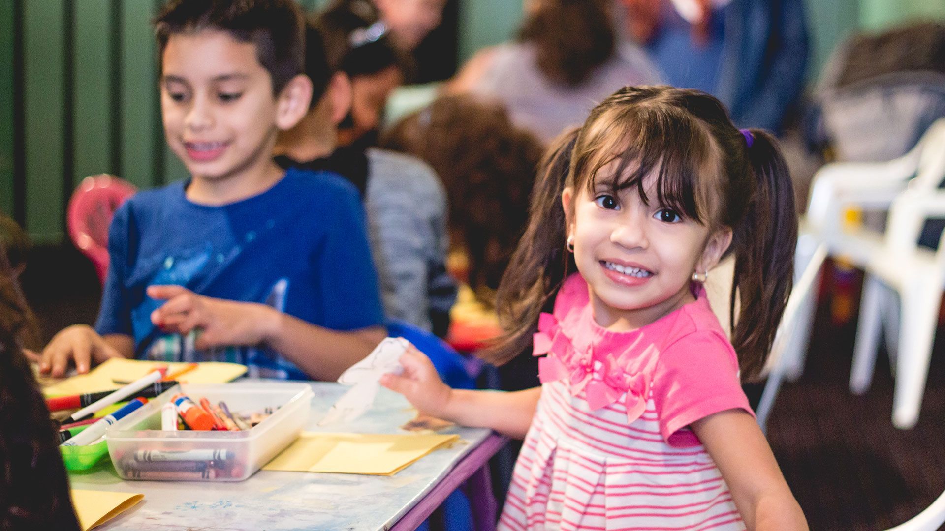 Two children smiling at a craft table, with crayons and paper in a colorful indoor setting