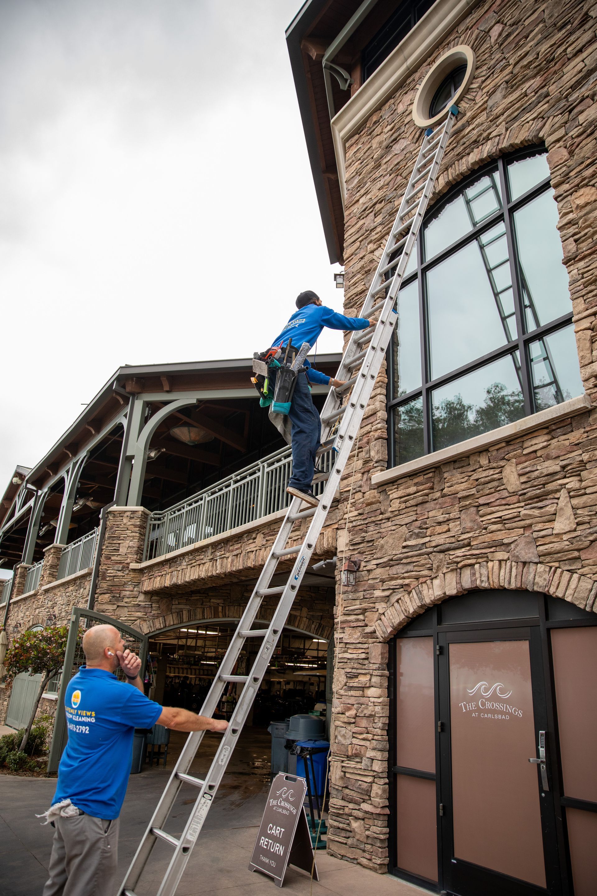 Workers on ladders repairing a brick building’s exterior near a large window.
