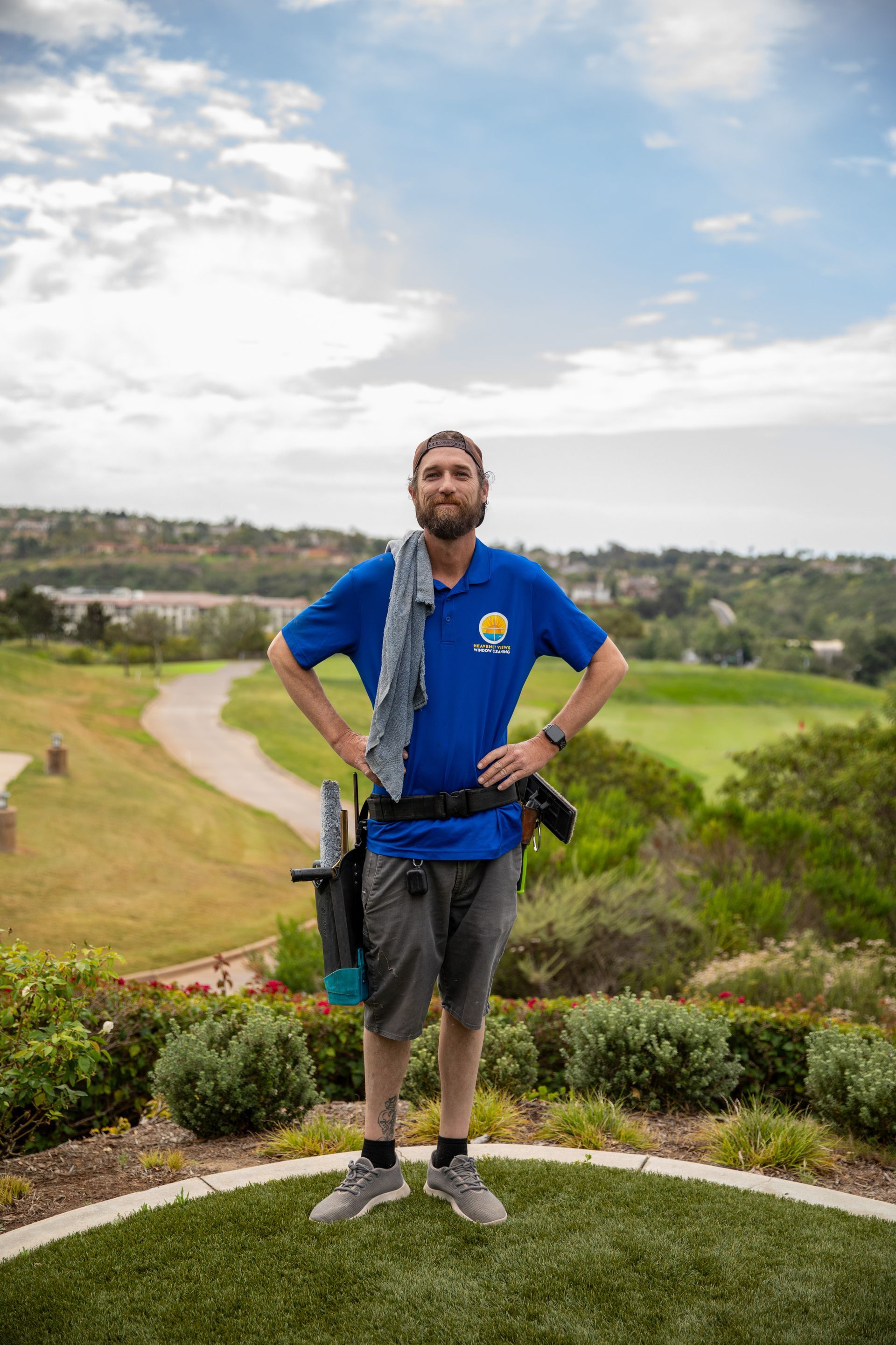 Man standing on a golf course hill with hands on hips, wearing a blue jacket and carrying golf clubs.