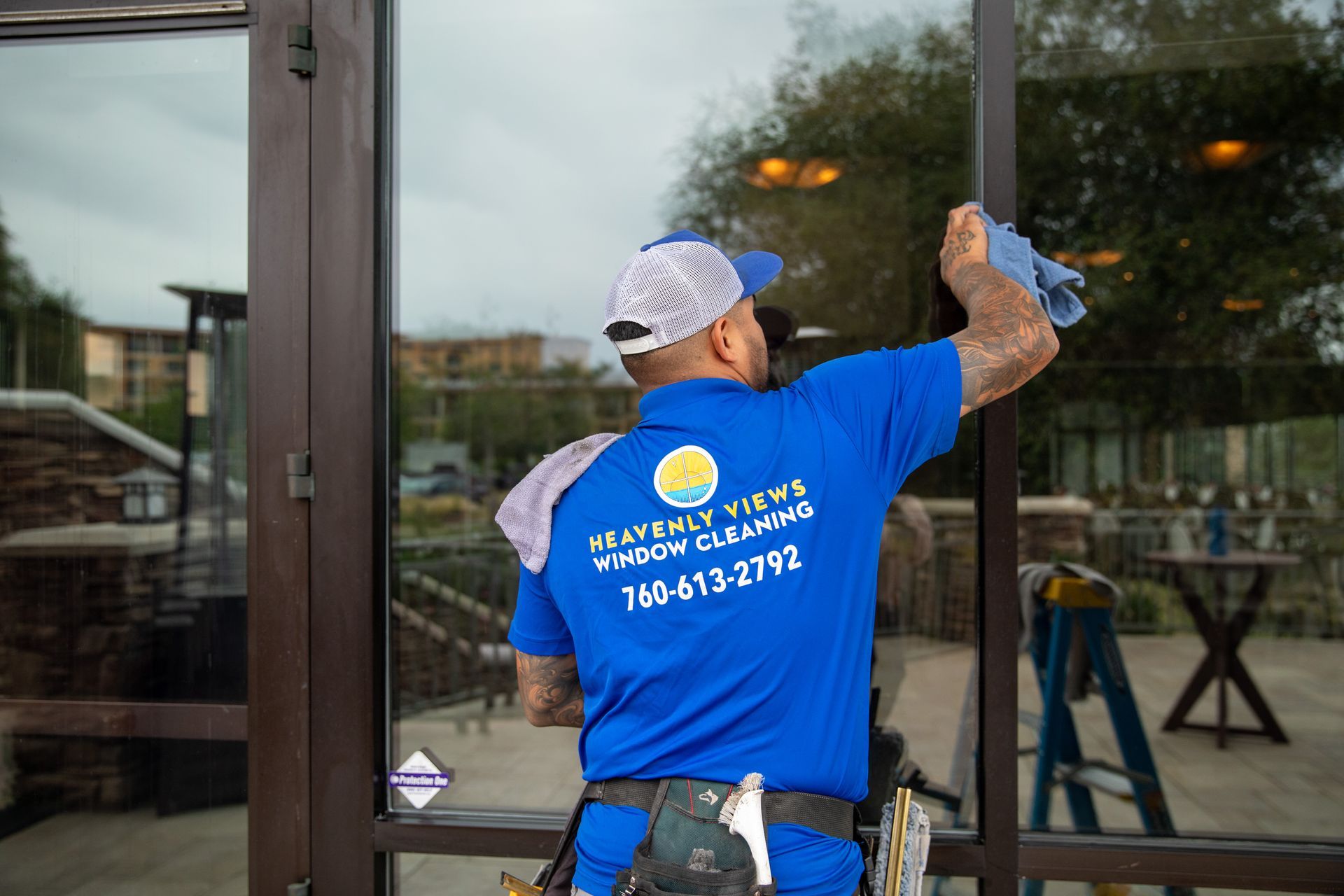 Worker in blue shirt cleaning a glass storefront with a squeegee and ladder nearby